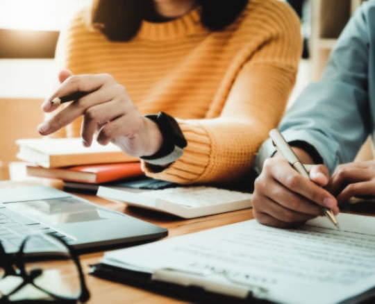 Emphasis is on the yellow-shirted consultant holding a pencil and the business owner holding a pen reading the terms of a joint venture contract with a partner company